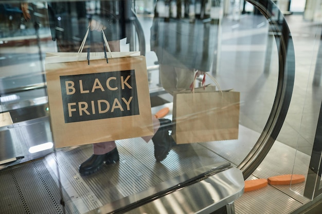 Captura recortada de una mujer irreconocible sosteniendo bolsas de compras con el cartel de venta del Viernes Negro mientras estaba de pie en la escalera mecánica en el centro comercial, copiando espacio