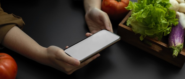 Captura recortada de mujer mirando receta en smartphone de pantalla en blanco horizontal mientras se preparan ingredientes de verduras frescas