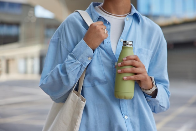 Foto captura recortada de una mujer sin rostro desconocida que usa camisa azul lleva una bolsa de tela sostiene una botella con paseos de agua dulce en la calle contra un fondo borroso siente poses sedientas contra el entorno urbano
