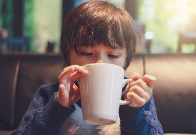 Captura recortada de niño bebiendo chocolate caliente en el café con tono cálido, niño niño sano soplando bebida caliente en la cafetería en invierno.