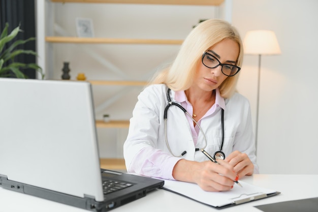 Captura de retrato de una doctora de mediana edad sentada en el escritorio y trabajando en el consultorio médico