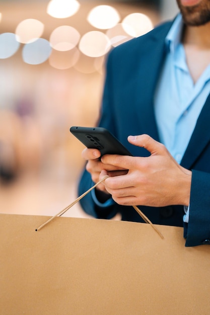 Foto captura vertical recortada de un joven irreconocible con traje de moda usando un teléfono móvil parado en el pasillo del centro comercial, sosteniendo bolsas de papel con compras, fondo borroso, primer plano