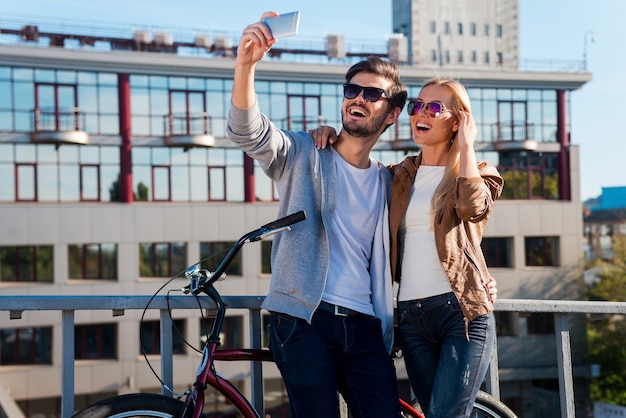 Capturando amor e diversão. Lindo casal jovem fazendo selfie em seu telefone inteligente e sorrindo em pé perto de uma bicicleta ao ar livre