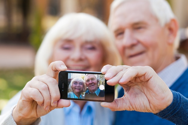 Capturando el amor sin fin. Feliz pareja senior uniéndose entre sí y haciendo selfie mientras está de pie al aire libre