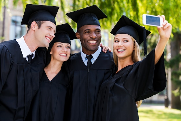 Capturando un momento feliz. Cuatro graduados universitarios en togas de graduación de pie cerca uno del otro y haciendo selfie