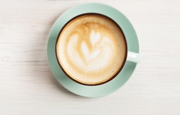 Capuchino con espuma espumosa, primer plano de la vista superior de la taza de café azul en la mesa de madera blanca. Café y bar, concepto de arte barista.