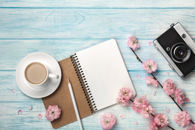 Capuchino de taza blanca con flores de sakura, cuaderno y cámara de fotos antigua en una mesa de madera azul