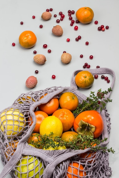 Caqui, manzanas y limón en bolsa de malla reutilizable. Lichi, arándanos y mandarinas en la mesa. Fondo gris. Vista superior