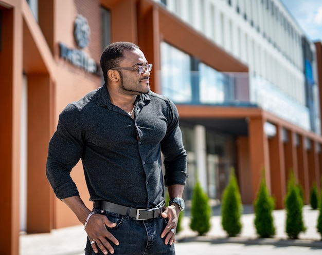 Cara africano com bíceps grande em pé na rua segurando as mãos nos quadris Homem de camisa apertada mostrando seu bíceps enorme Macho em frente ao centro de negócios, olhando para a frente
