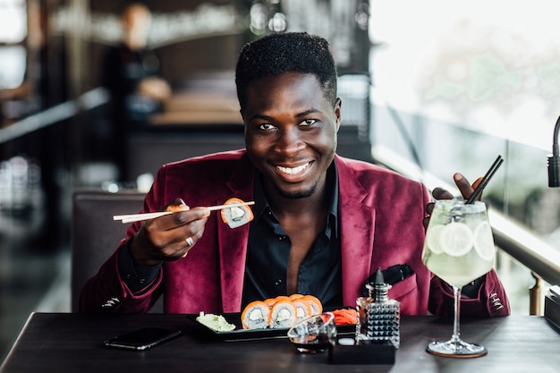 Cara africano de cabelo encaracolado alegre segurando rolos de sushi de pauzinhos. terraço do restaurante de peixes de comida chinesa. mojito na mesa.