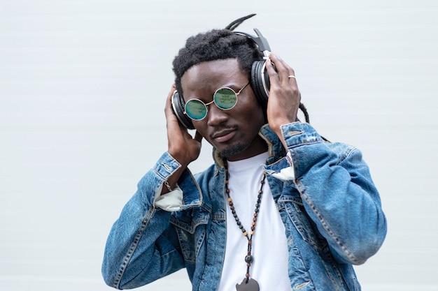 Foto cara africano elegante em jeans com dreadlocks e óculos de sol ouve música em fones de ouvido em um branco