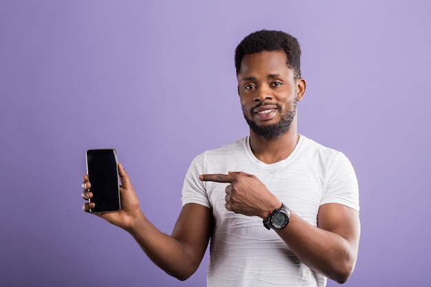 Cara africano feliz positivo vestido com roupa casual, mostrando o smartphone preto moderno e apontando para o dispositivo com o dedo indicador, sorrindo sobre a parede violeta do estúdio. tecnologia, conceito de comunicação.