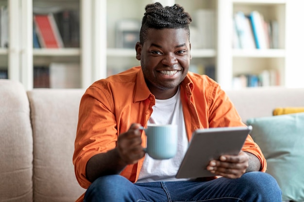 Cara africano positivo relaxando em casa segurando o tablet e a caneca