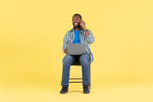 Cara afro-americana usando laptop falando no celular fundo amarelo