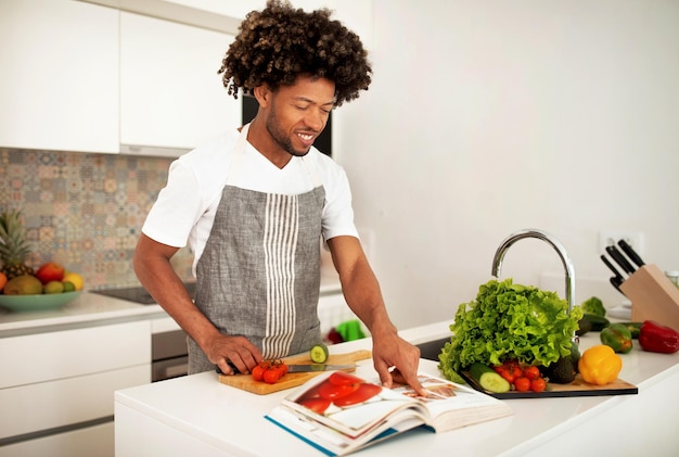 Cara afro-americano sorridente lendo livro culinário de papel na cozinha