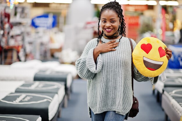 Cara amor corazón emoji. Mujer africana con almohada en una tienda de muebles para el hogar moderno.