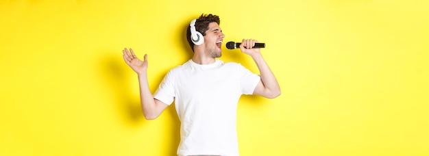 Cara apaixonado em fones de ouvido segurando microfone cantando música de karaokê em pé sobre fundo amarelo