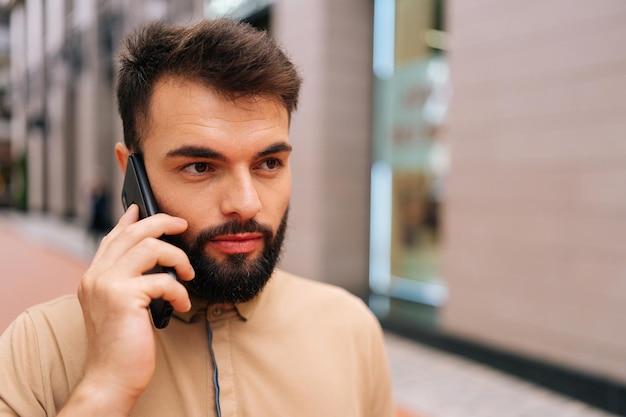 Cara aproximada de um jovem barbudo bonito andando na rua da cidade e falando em smartphone no fundo do prédio