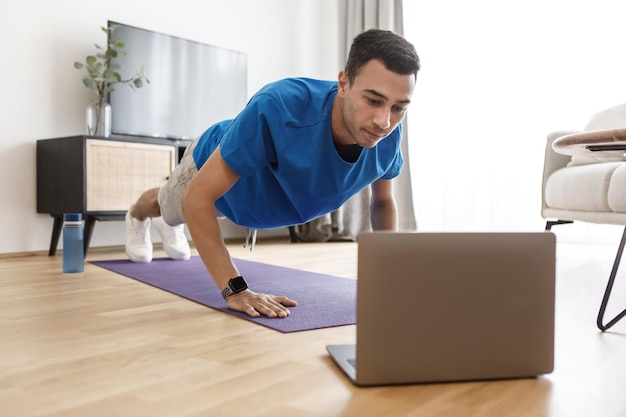 Cara árabe bonito em roupas esportivas fazendo flexões tendo aula de fitness online via laptop do espaço de cópia em casa