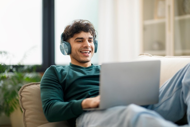 Cara árabe digitando e navegando na web no laptop usando fones de ouvido dentro de casa