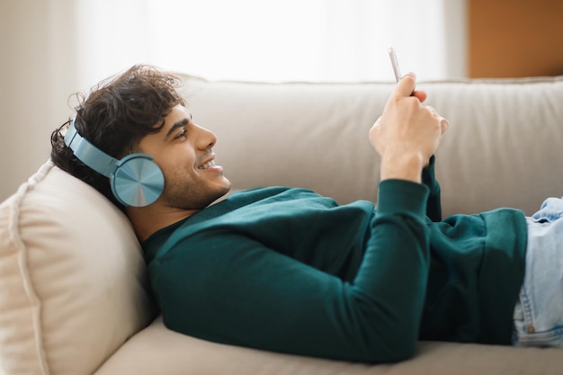 Cara árabe feliz usando fones de ouvido usando celular relaxando em casa