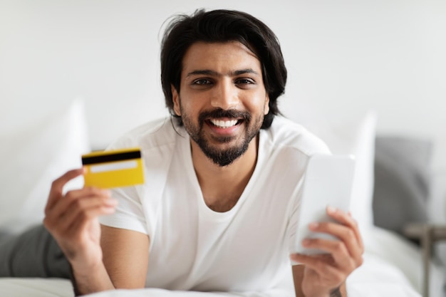 Cara árabe milenar sorridente com barba deitada na cama mostra cartão de crédito e smartphone à luz
