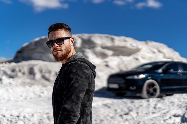 Cara atraente em roupas da moda com carro de luxo e fundo de pedras brancas. homem elegante modelo em óculos de sol.