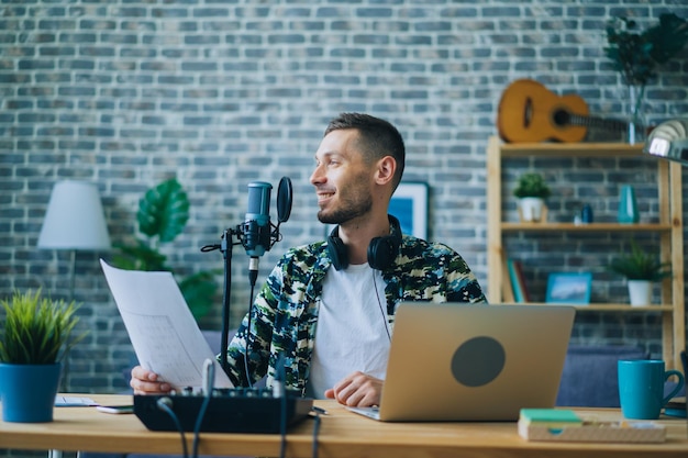 Cara atraente gravando podcast usando microfone segurando um pedaço de papel dentro de casa