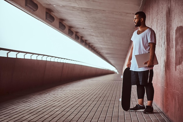 Cara barbudo afro-americano vestido com uma camisa branca e shorts esportivos segura em um laptop e skate enquanto está em uma passarela debaixo de uma ponte, olhando para longe.