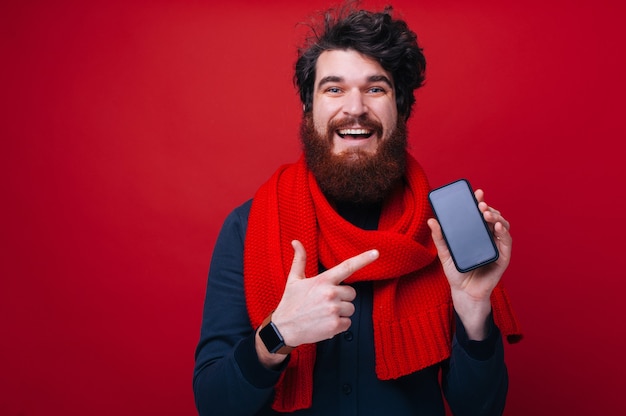 Cara barbudo bonito, usando um lenço vermelho, apontando para a tela do celular enquanto olha para a câmera, sobre um fundo isolado