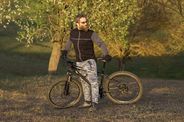 Cara barbudo brutal de esportes em uma bicicleta de montanha moderna Ciclista nas colinas verdes na primavera