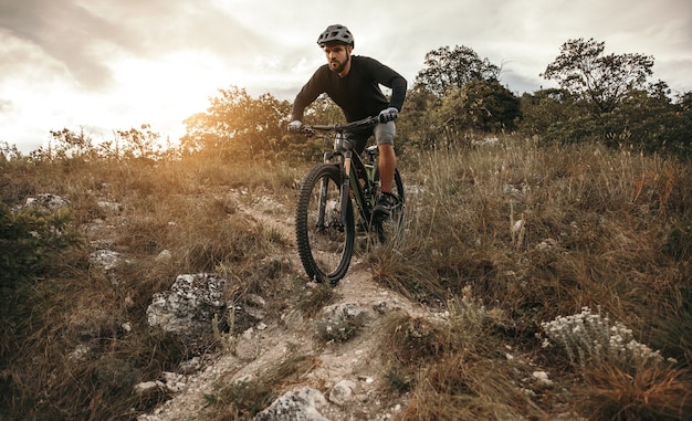 Cara barbudo de corpo inteiro andando de bicicleta moderna em uma colina gramada contra o céu nublado do pôr do sol no campo