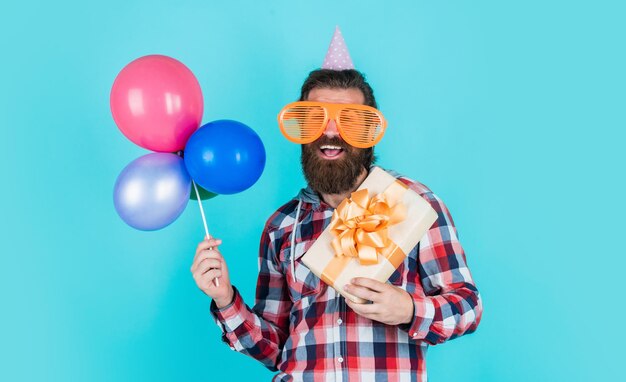 Cara barbudo elegante com penteado moderno usa camisa quadriculada segura balões de festa e caixa de presente, felicidade.