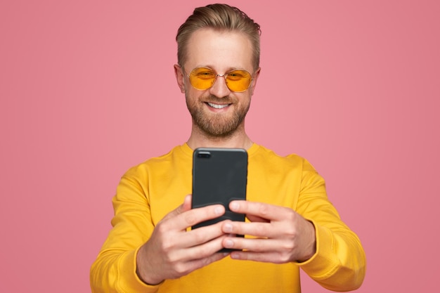 Cara barbudo hipster feliz em roupa amarela e óculos escuros se comunicando via celular e tirando uma selfie contra um fundo rosa