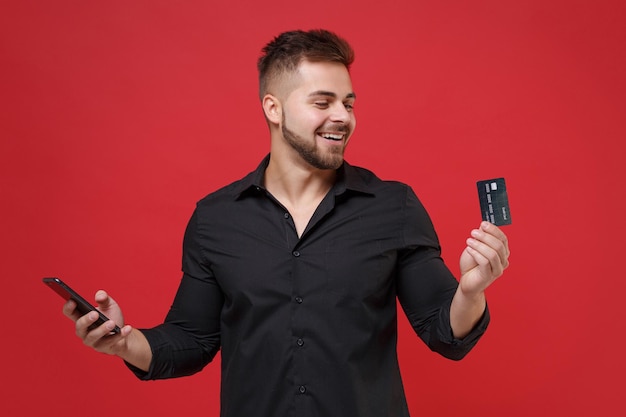 Cara barbudo jovem alegre na camisa preta clássica posando isolado no retrato de estúdio de fundo vermelho. Conceito de estilo de vida de emoções sinceras de pessoas. Mock up espaço de cópia. Segure o cartão de crédito, telefone celular.