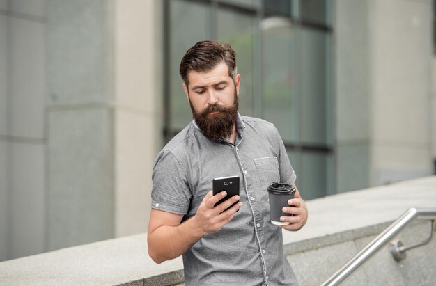 Cara barbudo navegando nas mídias sociais no celular enquanto bebe café ao ar livre Usando a Internet móvel na comunicação móvel do dispositivo móvel