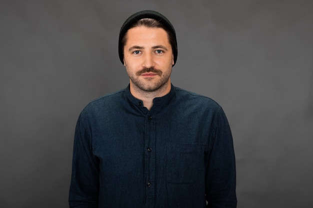 Cara bonito com a barba por fazer e boné de malha posando no cenário cinza do estúdio
