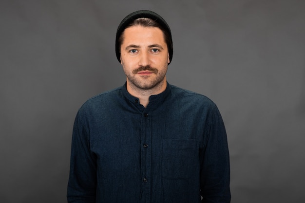 Foto cara bonito com a barba por fazer e boné de malha posando no cenário cinza do estúdio