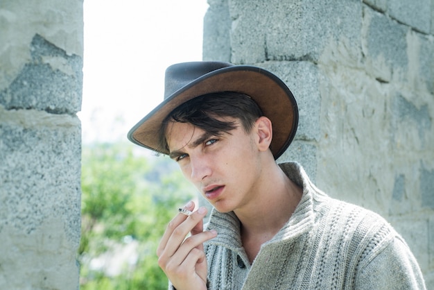 Cara bonito com rosto confiante. close-up jovem fumando um cigarro, charuto. homem bonito usa chapéu de cowboy.