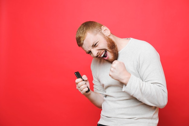 Cara bonito com smartphone moderno fazendo caretas e gesticulando com as duas mãos em um fundo vermelho brilhante