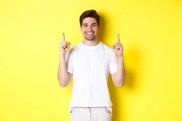 Foto cara bonito de camiseta branca apontando o dedo para cima, mostrando ofertas de compras, em pé sobre a parede amarela