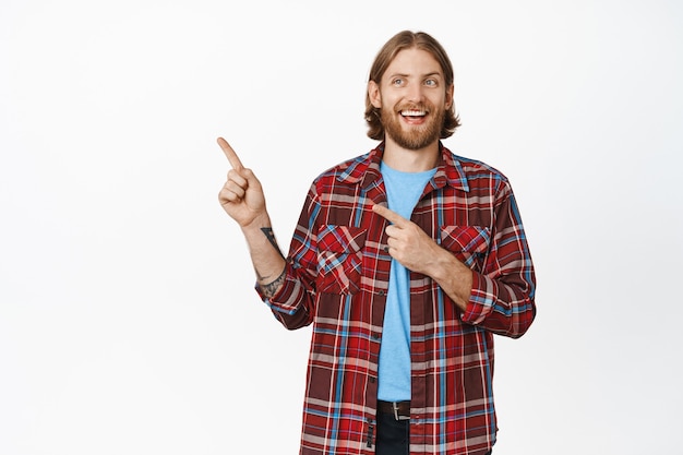 Cara bonito e sorridente hipster, homem loiro e branco com barba, olhando e apontando os dedos deixados no branco