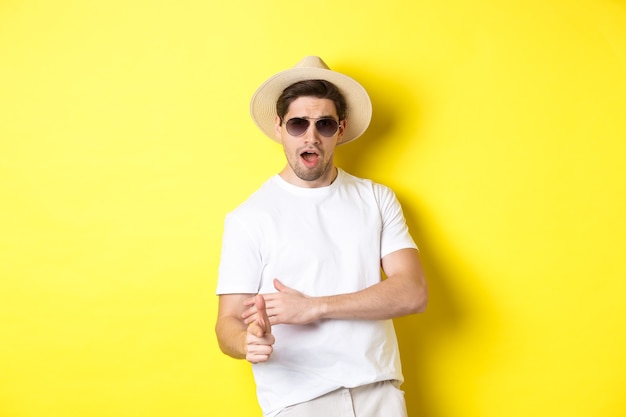 Cara bonito fazendo um tiro no dedo e parecendo atrevido, vestido para as férias de verão com chapéu de palha e