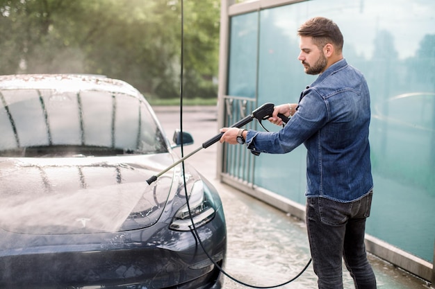 Cara bonito na camisa jeans lavando seu carro elétrico de luxo moderno enxaguando o sabão com jato de água de alta pressão Lavagem de carro manual com água pressurizada na lavagem de carro fora