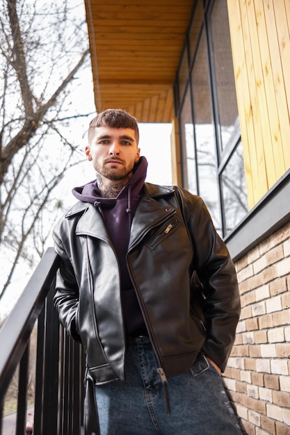 Cara bonito na moda brutal em roupas casuais da moda com uma jaqueta de couro do lado de fora da casa na rua Cara legal de moda hipster