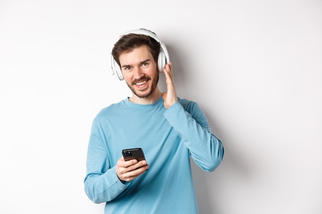 Cara bonito ouvindo música em fones de ouvido sem fio, sorrindo para a câmera e usando o smartphone, em pé sobre um fundo branco.