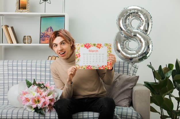 Cara bonito sorridente no feliz dia da mulher segurando o calendário sentado no sofá na sala de estar