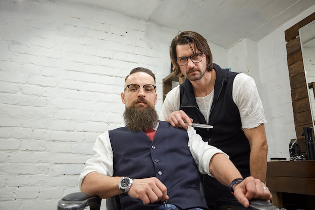 Cara brutal na barbearia moderna. cabeleireiro faz penteado um homem com uma barba longa. mestre cabeleireiro faz penteado por tesoura e pente