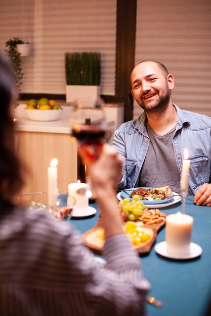 Cara celebrando relacionamento na cozinha com esposa feliz segurando um copo de vinho tinto, caminhando feliz sentado à mesa da sala de jantar, desfrutando da refeição em casa, tendo um tempo romântico à luz de velas