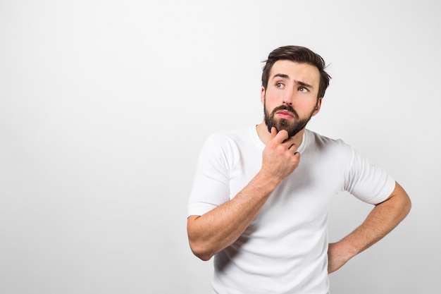 Cara com barba e pensativo está vestindo camisa branca e posando. ele está mantendo o braço direito perto do queixo e pensando. isolado na parede branca